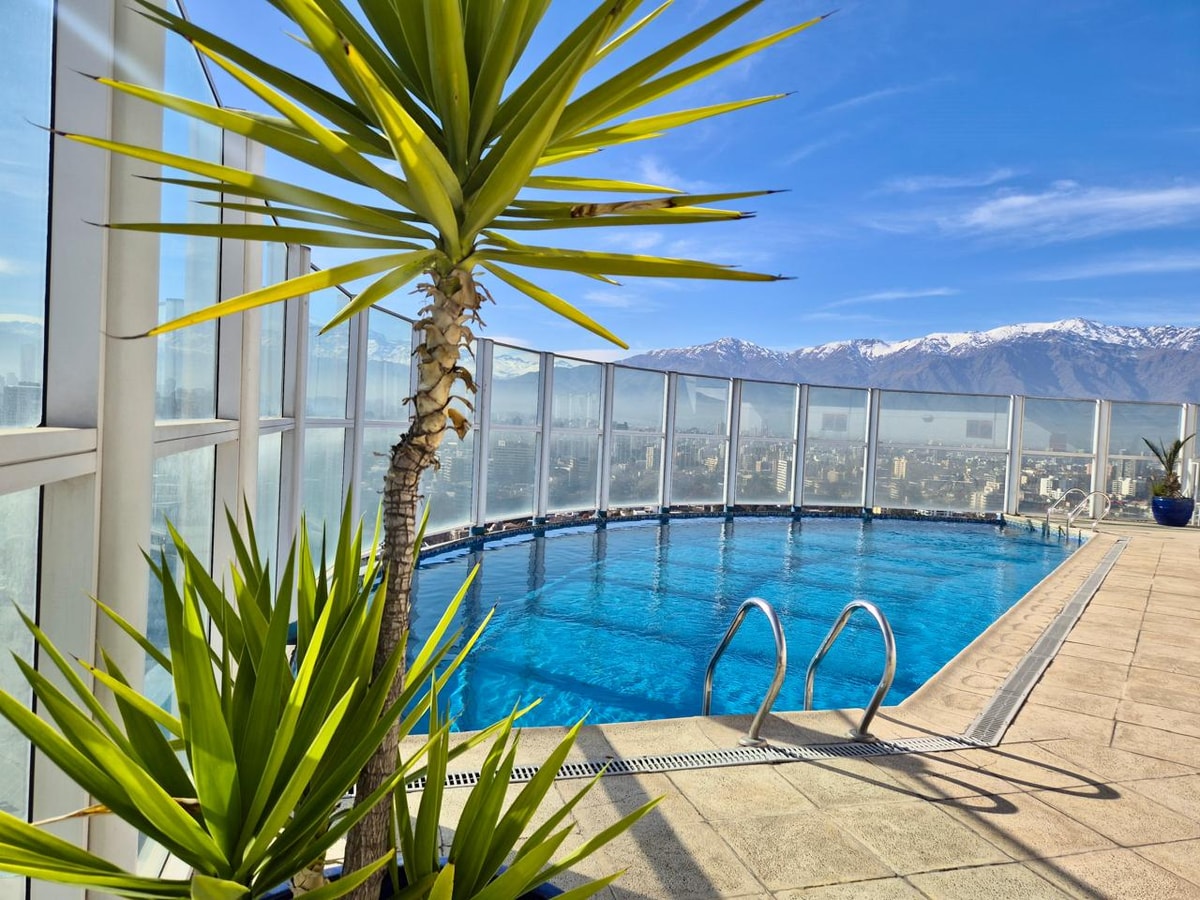 Piscina del edificio en la azotea, cerrada con paneles de vidrio, con solárium de baldosas y vista a la ciudad y la cordillera nevada
