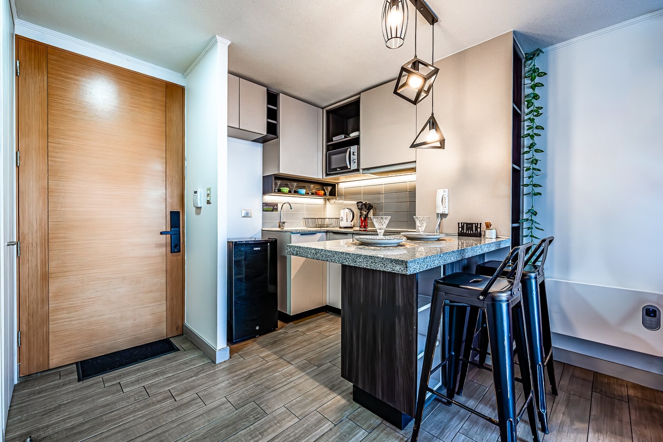 Cocina y living con vista a la puerta de entrada de madera, barra con banquetas y estante decorativo al fondo