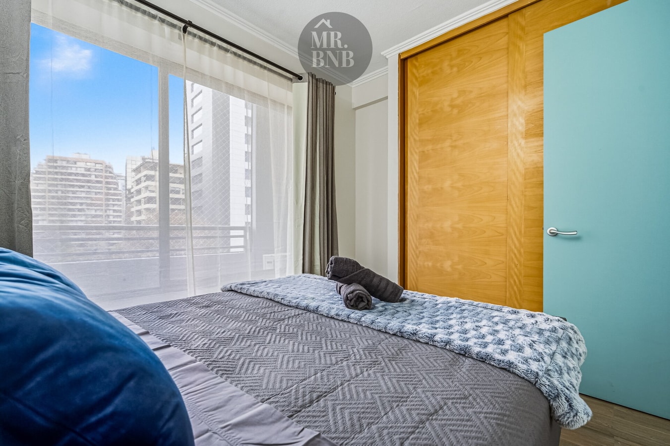 Dormitorio con cama, puerta celeste y ventanal con vista a edificios