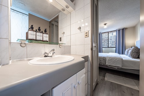 Baño con lavamanos y puerta abierta hacia un dormitorio con camas visibles al fondo.
