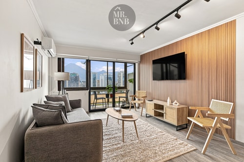 Living con sofá café, TV, sillón y ventanales con vista a la cordillera.