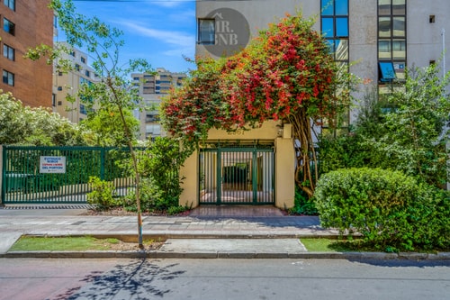Acceso y fachada del edificio, con reja, jardín y arbustos en flor junto a la vereda