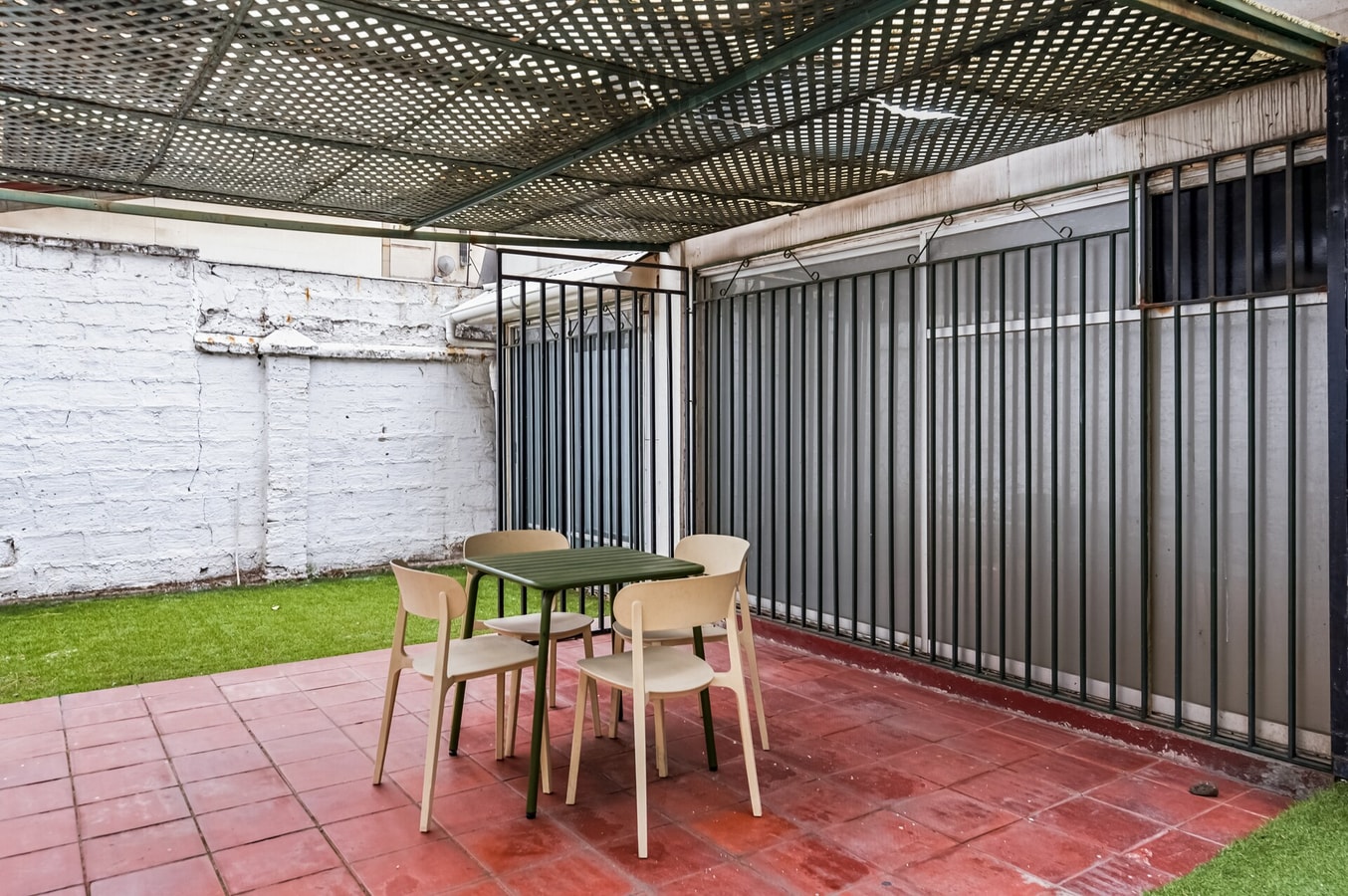 Patio trasero techado con estructura metálica, mesa, sillas, piso rojo y pasto sintético.