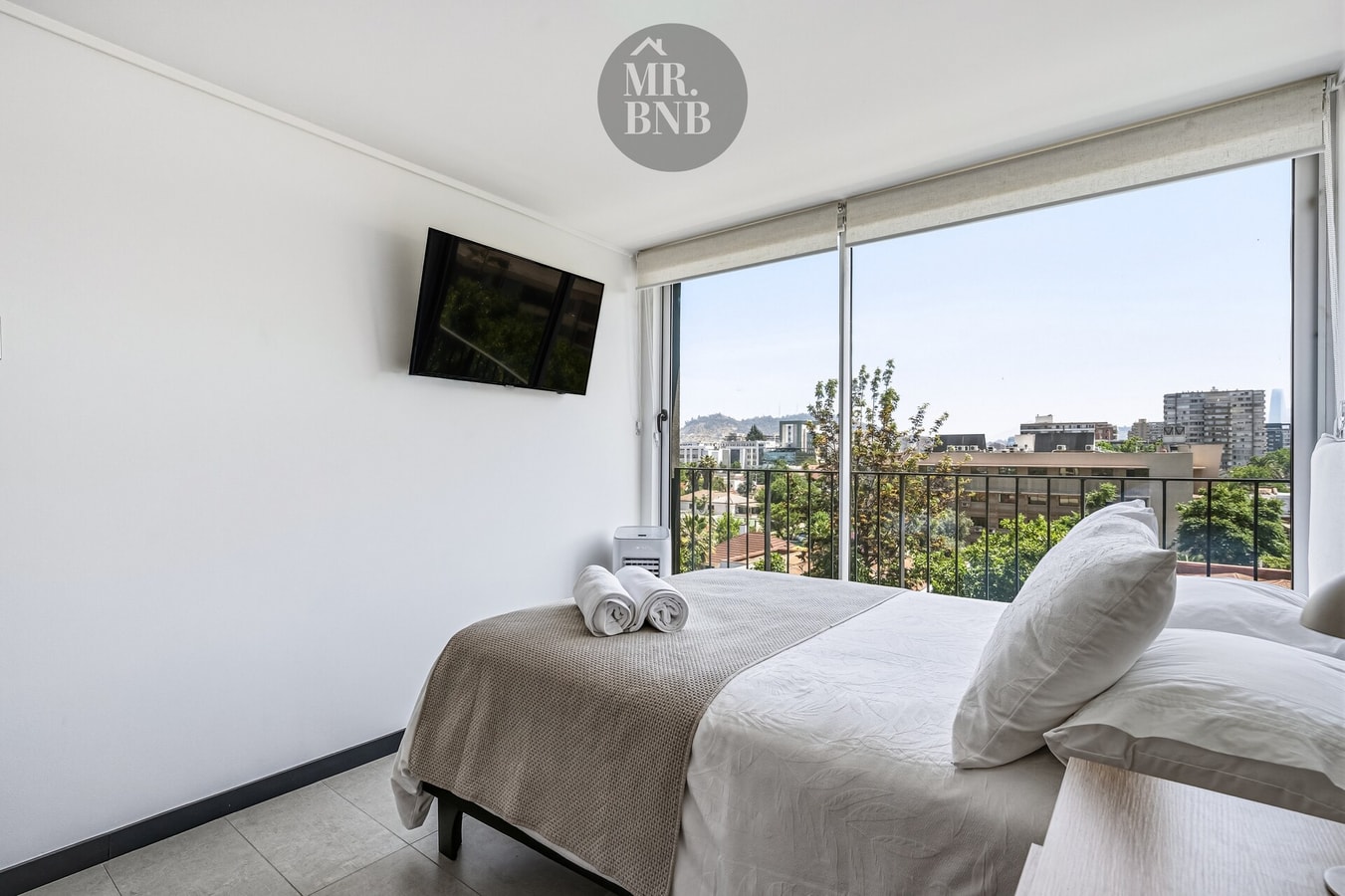 Dormitorio con cama, televisor y ventanal amplio con vista a edificios y cerros