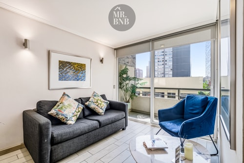 Living con sofá gris, sillón azul, cuadro y ventanal con vista a la ciudad.