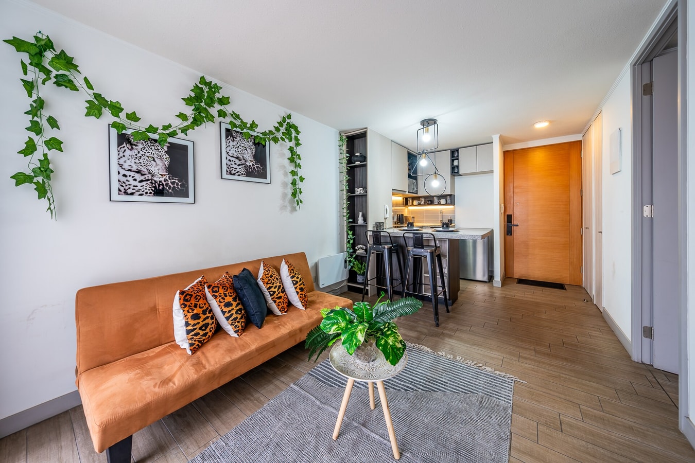 Living con sofá naranjo, cojines de estampado animal, enredadera artificial en la pared y barra de cocina con banquetas altas al fondo