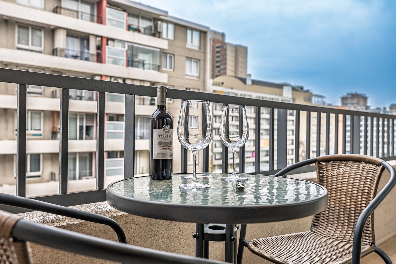 Terraza pequeña con mesa redonda y silla, vino y copas, con vista a edificios vecinos.