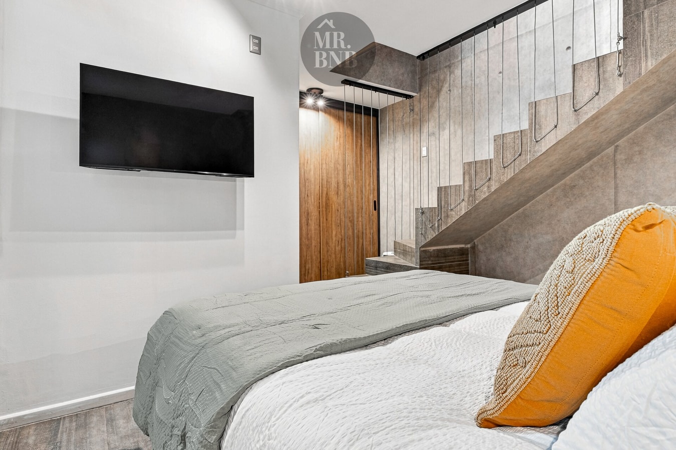 Dormitorio con cama, cojín mostaza, TV y escalera de hormigón