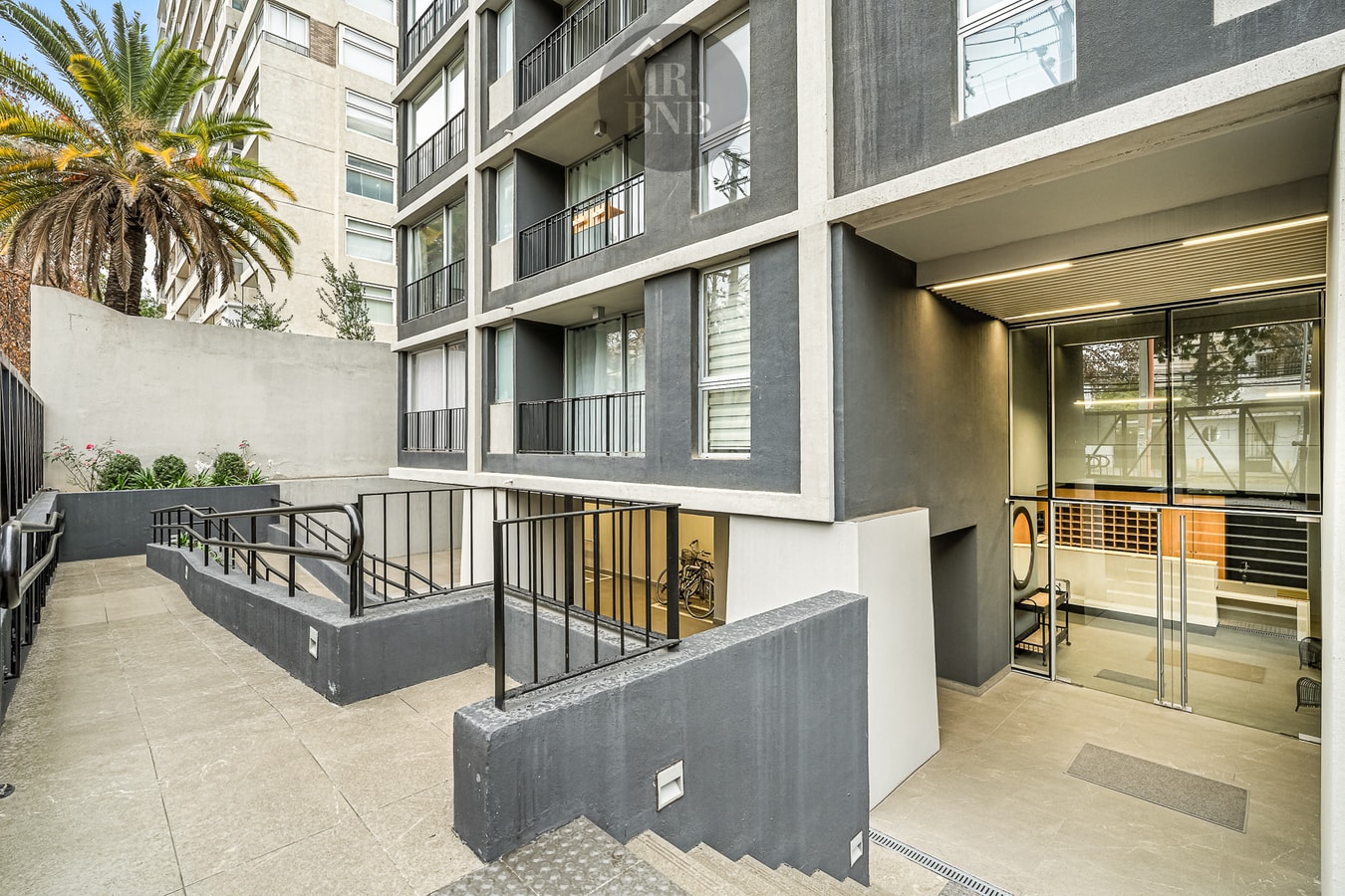 Patio de acceso del edificio visto desde la entrada, con escalera, palmera y la recepción vidriada al fondo